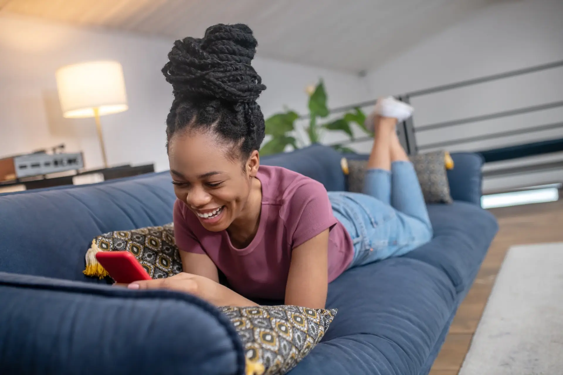 A Black woman scrolls through her phone lying on a plush navy sofa in a bright, cozy apartment